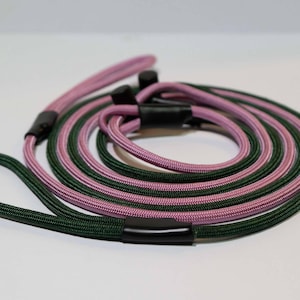 Pode incluir: Uma trela de corda bicolor com um cord&atilde;o entran&ccedil;ado verde-escuro e um cord&atilde;o entran&ccedil;ado roxo-claro. A trela inclui conectores de pl&aacute;stico preto e duas argolas integradas nas extremidades, enrolada numa forma circular sobre um fundo branco.