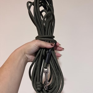 May include: A long olive green rope lead featuring a durable braided texture, a black heat-shrink tubing connector, and a polished silver-toned metal swivel snap hook at the end, coiled together for storage and portability.