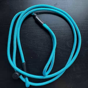 Pode incluir: Um cord&atilde;o turquesa brilhante feito de material de nylon tran&ccedil;ado, com uma textura lisa e uma se&ccedil;&atilde;o transversal circular. O cord&atilde;o &eacute; fixado com dois conectores cil&iacute;ndricos de pl&aacute;stico preto e uma pequena tampa preta, formando um la&ccedil;o.