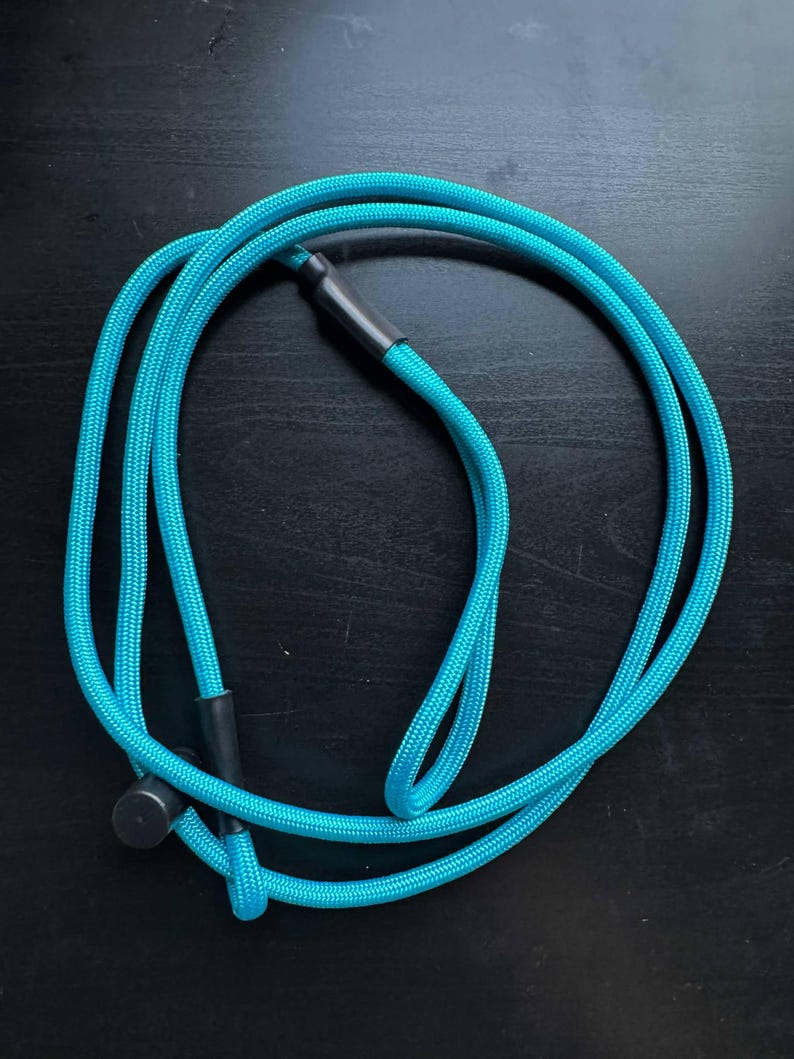 Pode incluir: Um cord&atilde;o turquesa brilhante feito de material de nylon tran&ccedil;ado, com uma textura lisa e uma se&ccedil;&atilde;o transversal circular. O cord&atilde;o &eacute; fixado com dois conectores cil&iacute;ndricos de pl&aacute;stico preto e uma pequena tampa preta, formando um la&ccedil;o.