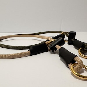 May include: A dual-coloured rope lead featuring a beige cord and an olive green cord. The ropes are secured with black cylindrical connectors and finished with polished gold-toned metal rings at each end for attachment points.