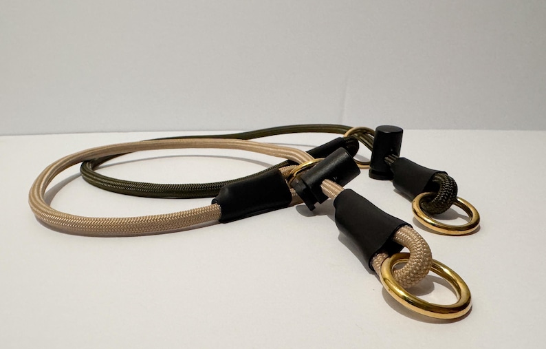 May include: A dual-coloured rope lead featuring a beige cord and an olive green cord. The ropes are secured with black cylindrical connectors and finished with polished gold-toned metal rings at each end for attachment points.