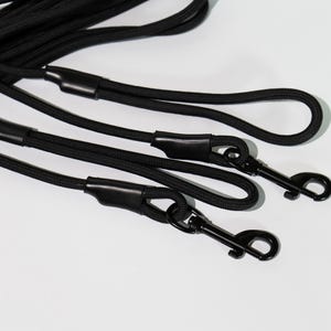 May include: A set of black rope leashes featuring durable braided cord construction. Each leash includes a sturdy black metal swivel snap hook at the end, secured with a smooth black rubberized sleeve to reinforce the connection point.