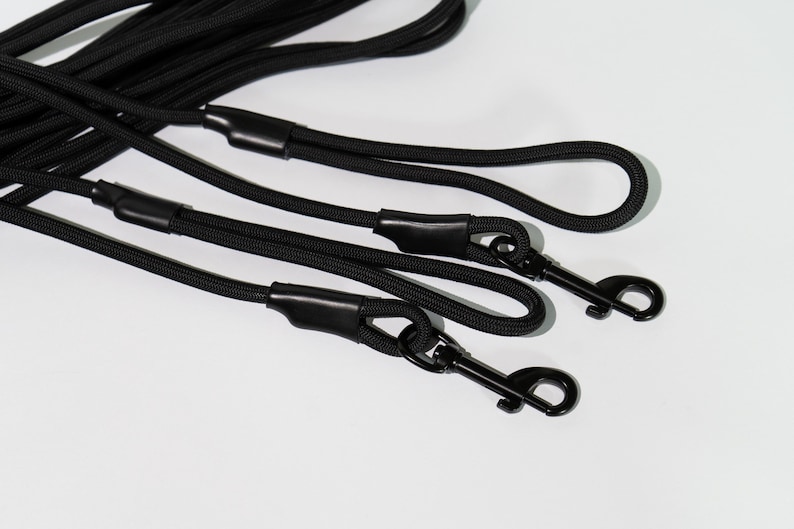May include: A set of black rope leashes featuring durable braided cord construction. Each leash includes a sturdy black metal swivel snap hook at the end, secured with a smooth black rubberized sleeve to reinforce the connection point.