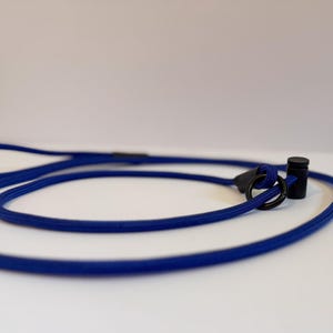 K&ouml;nnte beinhalten: Eine k&ouml;nigsblaue Seilleine mit einem schwarzen Metallring und einem schwarzen zylindrischen Stopper. Die Leine besteht aus einem strapazierf&auml;higen, gewebten Kordelmaterial mit einer Schlaufe an einem Ende und einem verschiebbaren Einstellmechanismus.