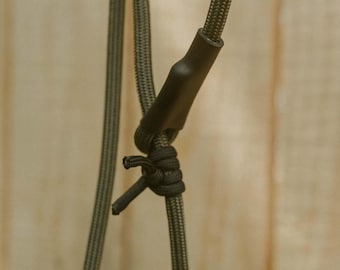 Könnte beinhalten: Ein olivgrünes Paracord-Lanyard mit einem schwarzen Schrumpfschlauch-Verbinder und einem sicheren schwarzen Knoten. Die Schnur hat eine texturierte, gewebte Oberfläche und ist vertikal angeordnet, wobei der Verbindungspunkt und das Knotendetail sichtbar sind.