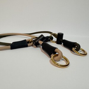May include: A dual-coloured rope lead featuring one beige and one olive green cord. Each cord is secured with black cylindrical connectors and matte black sleeves, finished with polished gold-toned metal rings at the ends for attachment.