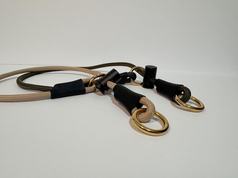 May include: A dual-coloured rope lead featuring one beige and one olive green cord. Each cord is secured with black cylindrical connectors and matte black sleeves, finished with polished gold-toned metal rings at the ends for attachment.