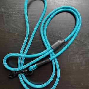 May include: A bright turquoise rope dog leash featuring a braided texture, black heat-shrink tubing accents, and a black metal swivel snap hook at the end. The leash is coiled in a loose loop, showcasing the vibrant blue cord and dark hardware.