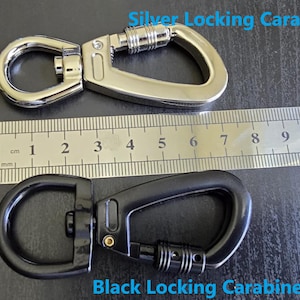 May include: A silver locking carabiner and a black locking carabiner positioned next to a metal ruler. The silver carabiner measures approximately 3 inches in length, while the black carabiner measures approximately 3 inches in length.