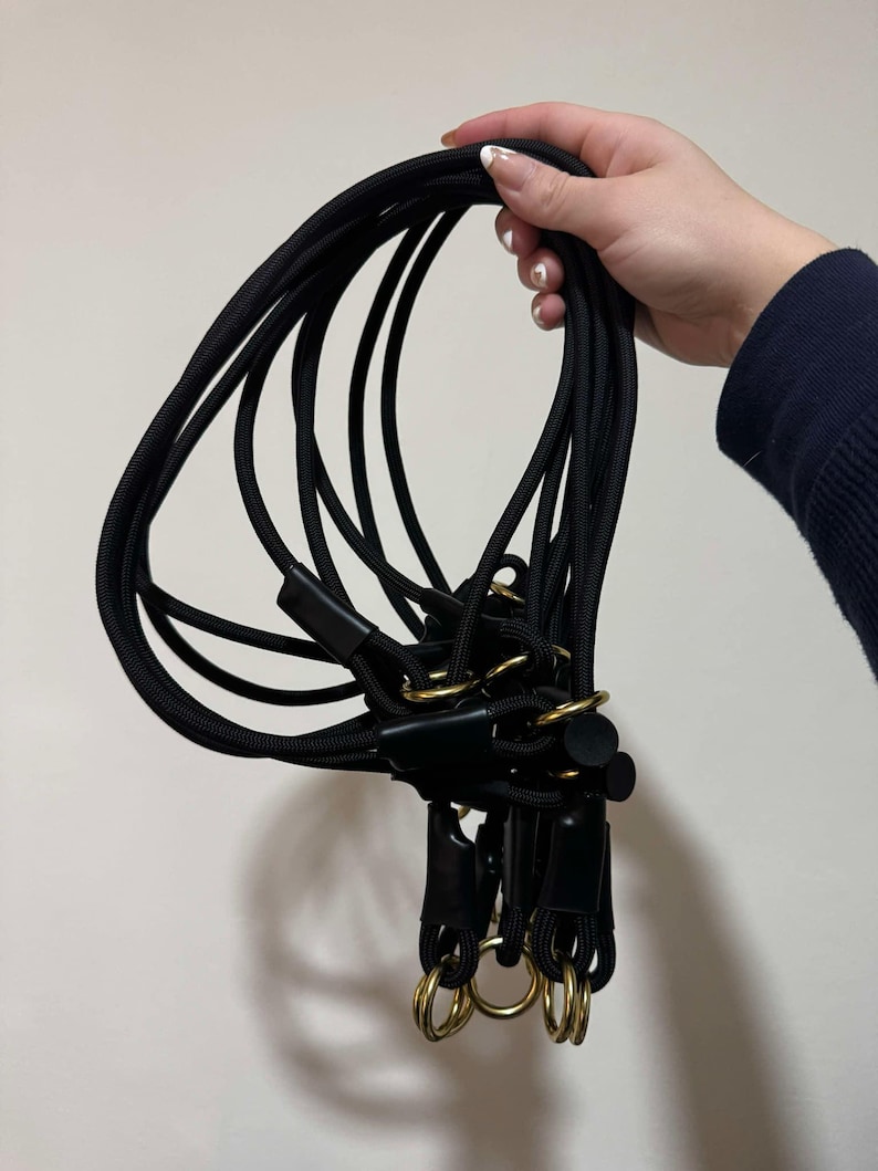 May include: A collection of black rope leads featuring gold-toned metal rings and black leather-like connectors. The ropes are flexible and looped, with multiple segments joined together by circular hardware, creating a functional design.