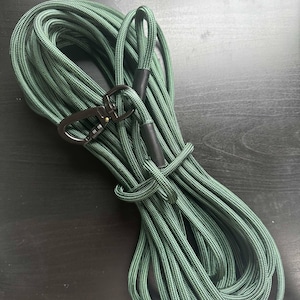 May include: A coiled forest green rope featuring a textured, braided surface. The rope includes a black metal swivel snap hook attached to one end and a black plastic sleeve covering the connection point, creating a secure and durable utility lead.
