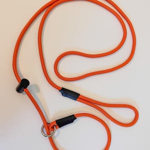 K&ouml;nnte beinhalten: Eine leuchtend orangefarbene Seilleine mit einem verschiebbaren schwarzen Kunststoffstopper, einem runden Metallring und zwei schwarzen Schrumpfschlauchverbindern. Das Seil hat eine strukturierte Oberfl&auml;che und bildet eine durchgehende Schlaufe.