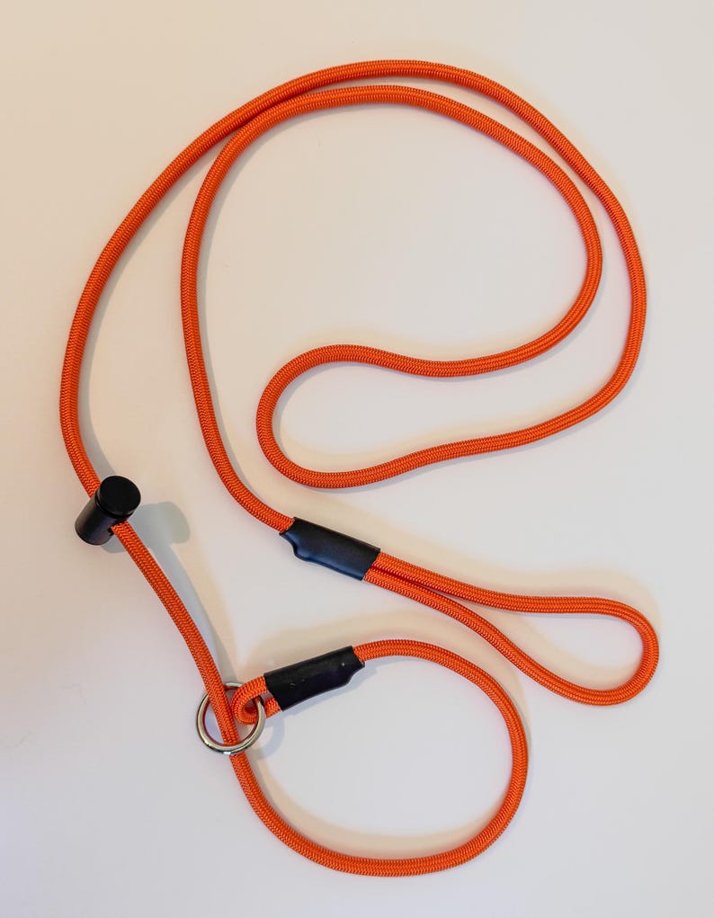 K&ouml;nnte beinhalten: Eine leuchtend orangefarbene Seilleine mit einem verschiebbaren schwarzen Kunststoffstopper, einem runden Metallring und zwei schwarzen Schrumpfschlauchverbindern. Das Seil hat eine strukturierte Oberfl&auml;che und bildet eine durchgehende Schlaufe.