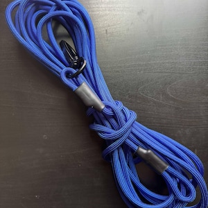 May include: A coiled length of bright blue braided rope featuring a black metal swivel clip at one end. The rope is secured with two black heat-shrink tubing sleeves that hold the loops in place, creating a durable and functional cord design.