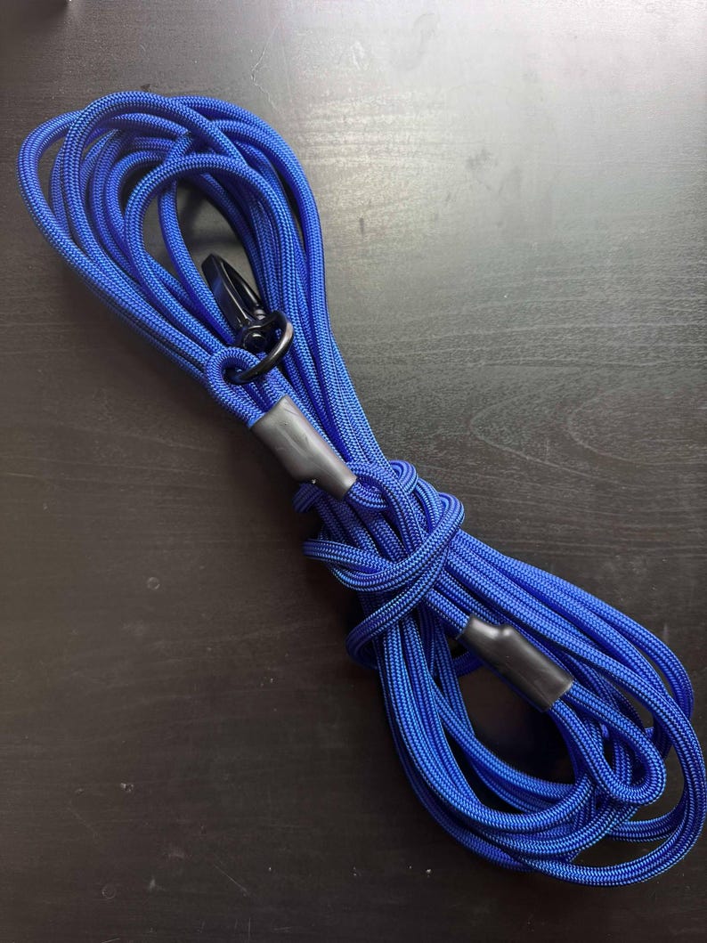 May include: A coiled length of bright blue braided rope featuring a black metal swivel clip at one end. The rope is secured with two black heat-shrink tubing sleeves that hold the loops in place, creating a durable and functional cord design.