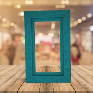 Könnte beinhalten: Ein türkisfarbener rechteckiger Bilderrahmen mit einem Blättermuster auf der Oberfläche des Rahmens. Der Rahmen steht auf einer Holzoberfläche. Der Hintergrund ist unscharf.