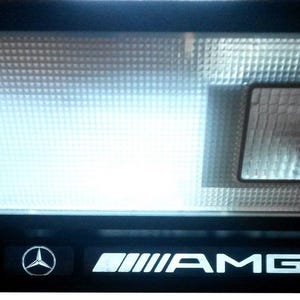 Può includere: Una luce interna per auto nera con una lampadina bianca e un logo AMG argentato.