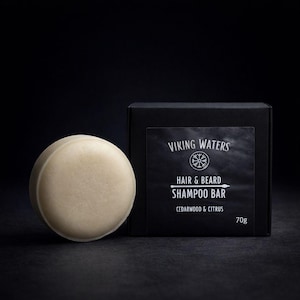 May include: A light beige, circular shampoo bar beside a black box. The box reads "VIKING WATERS HAIR & BEARD SHAMPOO BAR CEDARWOOD & CITRUS 70g". The shampoo bar is a solid, round shape, and the box is rectangular.
