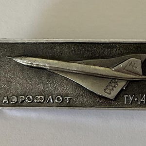 Pode incluir: Alfinete retangular prateado com um design estilizado de avião. O alfinete tem o texto cirílico "АЭРОФЛОТ" e "ТУ-144". O avião é retratado em perfil, com asas e cauda. O alfinete tem um fundo texturizado.