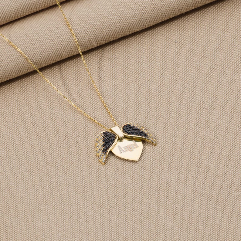 May include: A gold chain necklace with a heart-shaped pendant featuring black wings and the word "Angel" engraved on it.