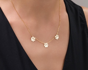 May include: A gold-colored necklace featuring three paw print charms. The necklace is worn against a black top, highlighting the delicate chain and the detailed paw print pendants. A charming accessory for pet lovers.