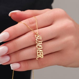 May include: A gold-colored necklace with the word "SUMMER" vertically displayed. The pendant is suspended from a delicate chain. The necklace is held by a person.