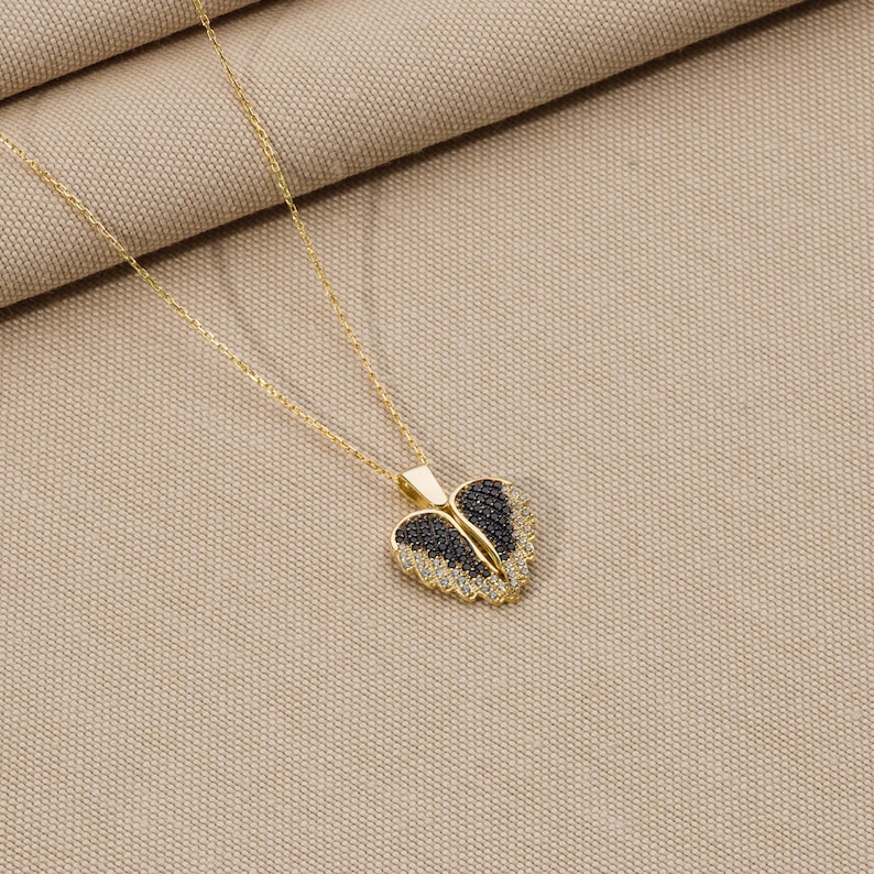 May include: A gold necklace with a heart-shaped pendant featuring black and white crystal accents. The pendant is shaped like a pair of angel wings.