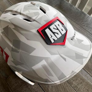 May include: White and gray camouflage baseball helmet with a red and black diamond-shaped patch that says "ASB" on it.