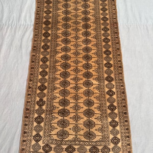 Puede incluir: Una alfombra rectangular tejida a mano con un diseño central de formas geométricas repetidas en tonos marrones y beige. La alfombra tiene un borde detallado y flecos en ambos extremos, mostrando un patrón tradicional.