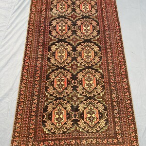 May include: A rectangular rug featuring a central pattern of repeating geometric shapes in black, brown, and peach. The rug has a detailed border with a repeating floral design. The rug is likely made of wool or a similar material.