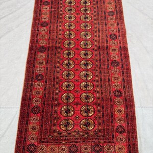 Puede incluir: Una alfombra rectangular roja con un patrón central de medallones dorados y negros repetidos. La alfombra tiene un borde detallado y flecos en los extremos. La alfombra es probablemente de lana y es adecuada para una sala de estar o un dormitorio.