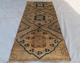 May include: A rectangular, tan-colored rug with a repeating geometric pattern in navy blue and brown. The rug features diamond and zigzag designs, with a fringed edge. The rug is placed on a white surface.