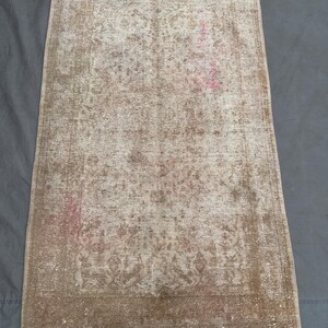 May include: A rectangular, distressed rug with a beige and brown color palette. The rug features a faded floral pattern and a white fringe along the shorter edge. The rug's design has a vintage, antique aesthetic.