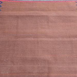 May include: A rectangular brown rug with a woven texture. The rug has a decorative border with a zigzag pattern in black, red, and blue. The rug is suitable for home decor.
