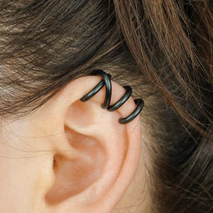 Könnte beinhalten: Nahaufnahme eines Ohrs, das mit einem schwarzen Ohrklemme und einem kleinen Perlenohrring geschmückt ist. Die Ohrklemme hat ein Spiraldesign, das sich um den oberen Ohrknorpel wickelt. Das Haar ist braun und zurückgebunden.