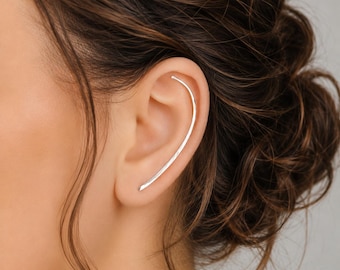 Puede incluir: Un pendiente tipo ear climber de plata con un diseño de barra larga, delgada y curva que sigue el contorno natural del borde exterior de la oreja. El metal tiene un acabado liso y pulido que refleja la luz, creando un aspecto limpio.