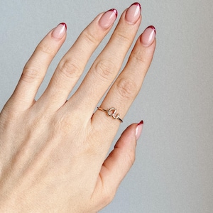 Puede incluir: Un anillo dorado con las letras "au" se muestra en una mano con uñas manicuradas. Las uñas tienen una base transparente con puntas rojas. El anillo es delicado y se lleva en el dedo anular.