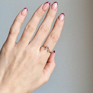 May include: A gold-colored wire ring in a cursive letter design is worn on a hand with manicured nails. The nails have a clear base with red glitter tips. The ring is the focal point, set against a neutral gray background.