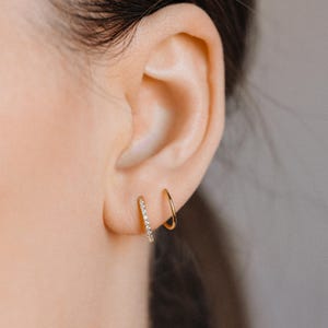 Pode incluir: Dois brincos de argola dourados usados no lóbulo da orelha. Uma argola é uma banda de ouro polido lisa, enquanto a outra é uma argola de ouro mais fina incrustada com uma fileira de pequenas pedras claras e brilhantes na borda externa.