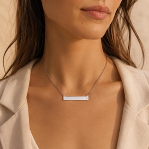 Könnte beinhalten: Eine silberfarbene horizontale Stabkette mit einer feinen Kette und einem einzelnen kleinen eingebetteten Stein auf der rechten Seite des rechteckigen Anhängers. Das Metall hat eine polierte Oberfläche und liegt am Schlüsselbein.