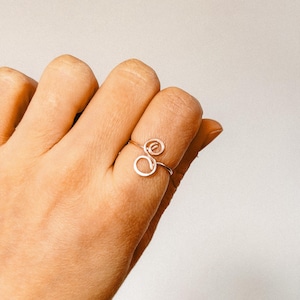 Könnte beinhalten: Ein filigraner, silberfarbener Drahtring mit einem Spiral- und Kreisdesign. Der Ring wird an einem Finger getragen und unterstreicht seinen minimalistischen und künstlerischen Stil. Das Design des Rings ist schlicht, aber elegant und für den Alltag geeignet.