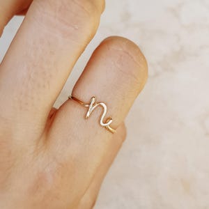 May include: A delicate gold-colored ring featuring the cursive letter "n". The ring is crafted from thin wire and sits on a finger. The ring's design is simple and elegant, suitable for everyday wear.