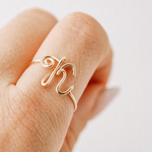 May include: A gold-colored wire ring in the shape of the letter "K" is displayed on a finger. The ring is crafted from a thin, continuous wire, forming the cursive letter with elegant loops and curves. The ring is a delicate piece of jewelry.
