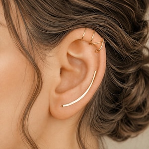 May include: A gold-toned ear cuff featuring three thin, stacked bands at the top of the ear cartilage and a single, smooth, curved ear climber bar extending along the lower lobe, creating a layered and minimalist appearance on the ear.