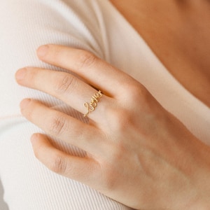 Può includere: Un delicato anello d'oro con la parola "love" scritta in corsivo. L'anello ha un design continuo, con le lettere collegate. Viene indossato su un dito, evidenziando la sua estetica semplice ed elegante. L'anello è d'oro.