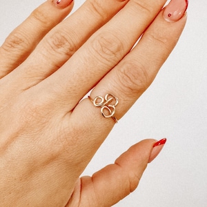 May include: A gold-coloured wire ring featuring the cursive letter "B" on a finger. The ring is delicate and minimalist, with a simple band. The background is a light, neutral colour.