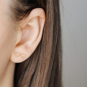 Könnte beinhalten: Nahaufnahme eines Ohrs mit einem kleinen, goldfarbenen Ohrring mit Unendlichkeitszeichen. Der Ohrring hat ein schlichtes, minimalistisches Design. Das Haar der Person ist braun und der Hintergrund ist ein neutrales Grau.