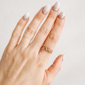 Puede incluir: Un anillo dorado con la inscripción "f&e" se lleva en un dedo. La mano tiene uñas manicuradas con esmalte blanco y puntas con purpurina plateada. El anillo es el punto focal sobre un fondo neutro.