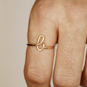 Può includere: Un anello color oro a forma di lettera "b" è mostrato su un dito. L'anello è delicato e sembra essere fatto di filo sottile. Lo sfondo è di un colore morbido e neutro, che mette in risalto il design dell'anello.